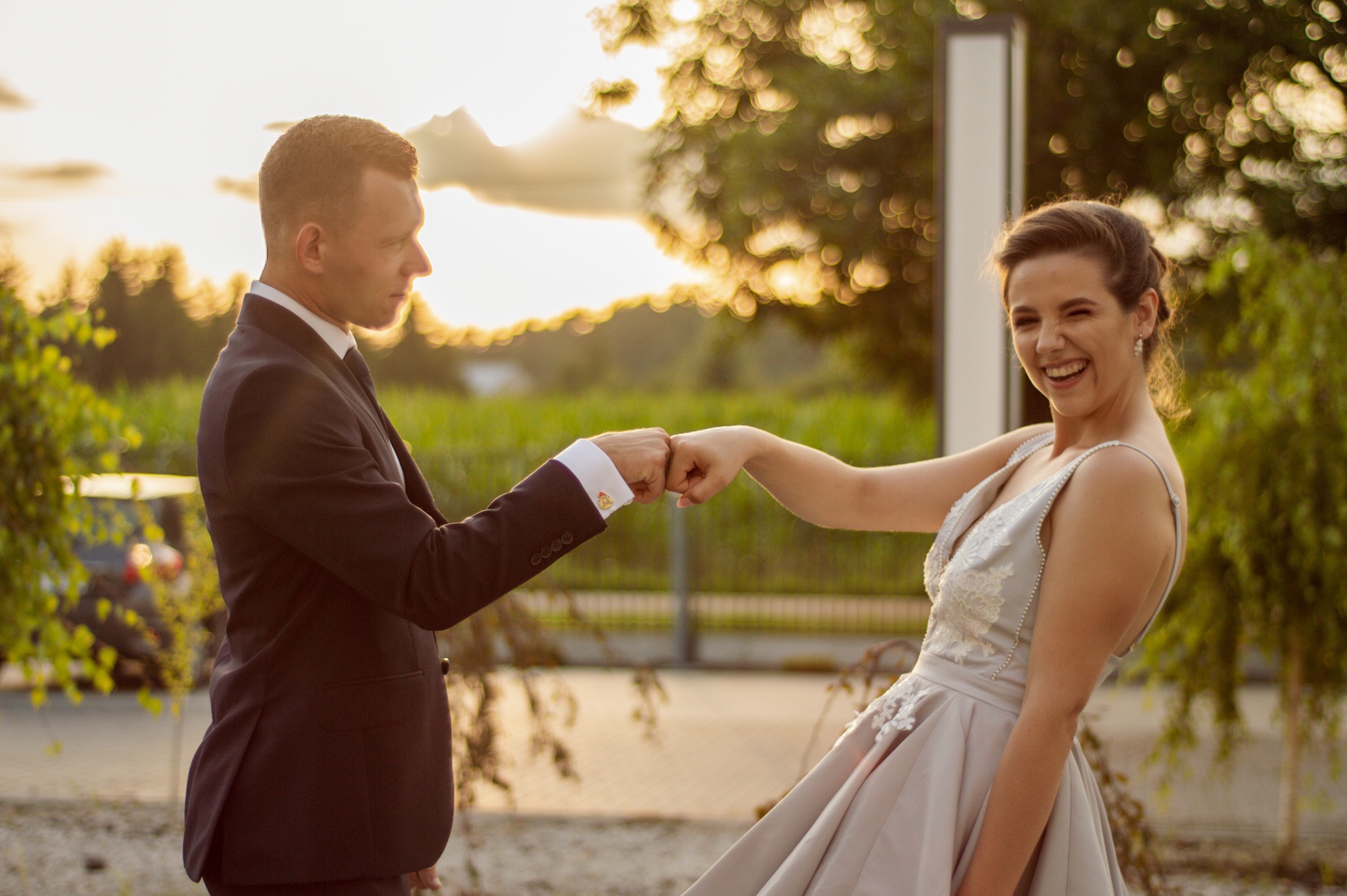 Uśmiechnięta panna młoda w sukni ślubnej i pan młody w garniturze witają się pięściami na tle zachodzącego słońca.