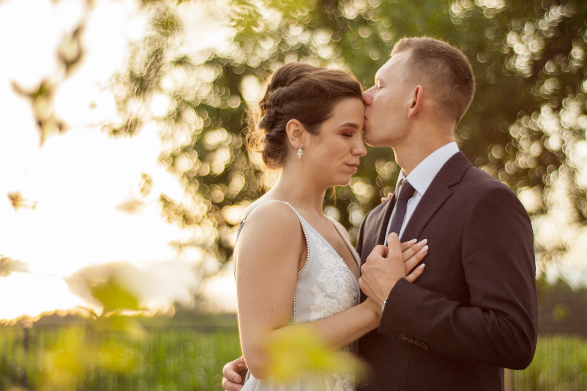 Panna młoda w sukni z koronkowym zdobieniem i diademem, obejmowana przez pana młodego w ciemnym garniturze, który całuje ją w czoło na tle rozmytych drzew i zachodzącego słońca.