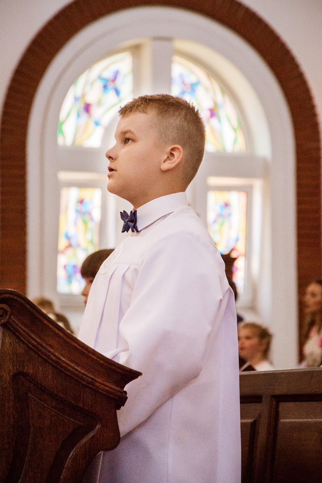 Chłopiec w albie komunijnej z granatową muchą, stojący przy drewnianej ambonie w kościele, w tle witrażowe okno.