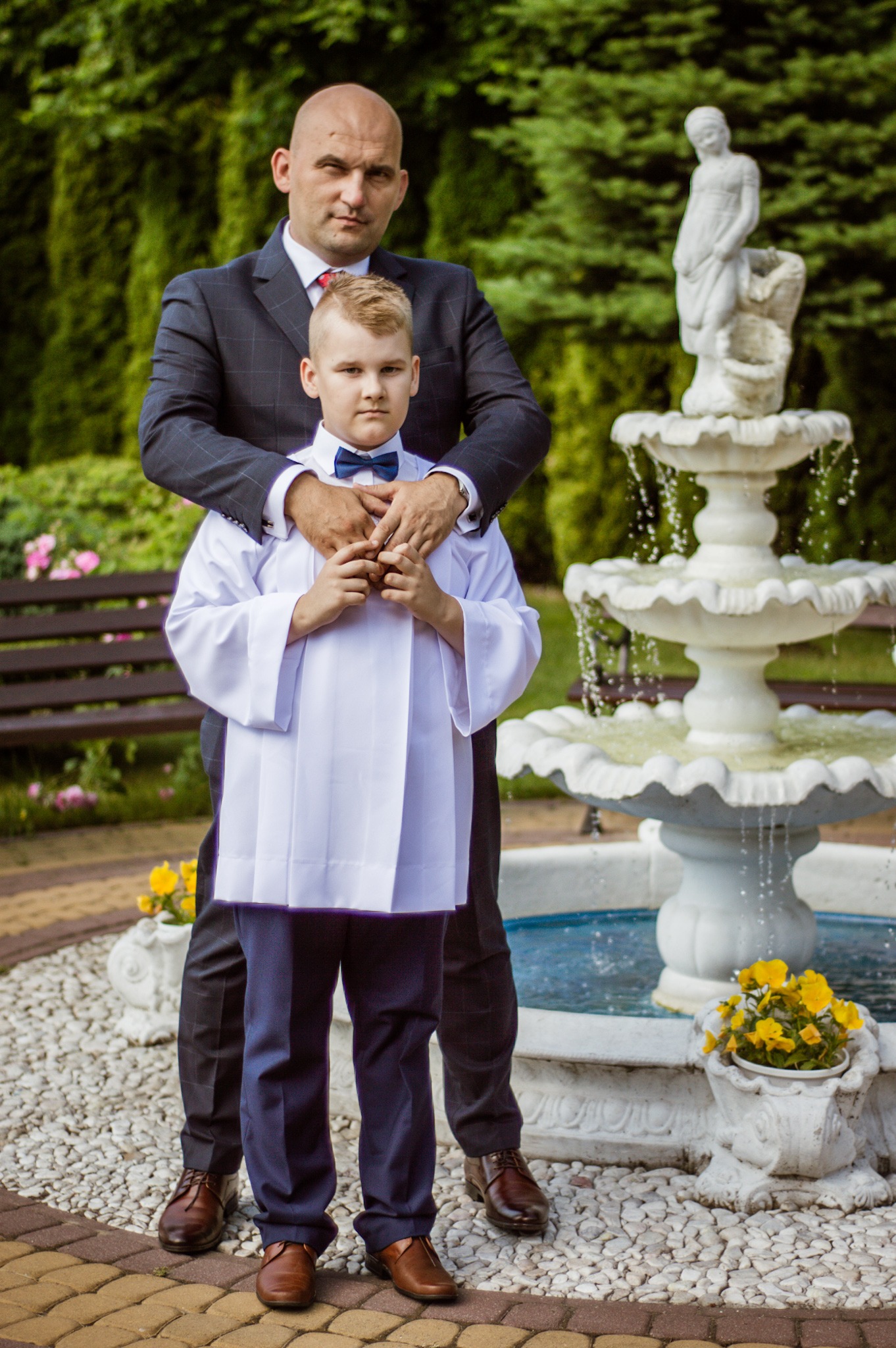 Chłopiec w stroju komunijnym i mężczyzna w garniturze pozują na tle fontanny w ogrodzie.