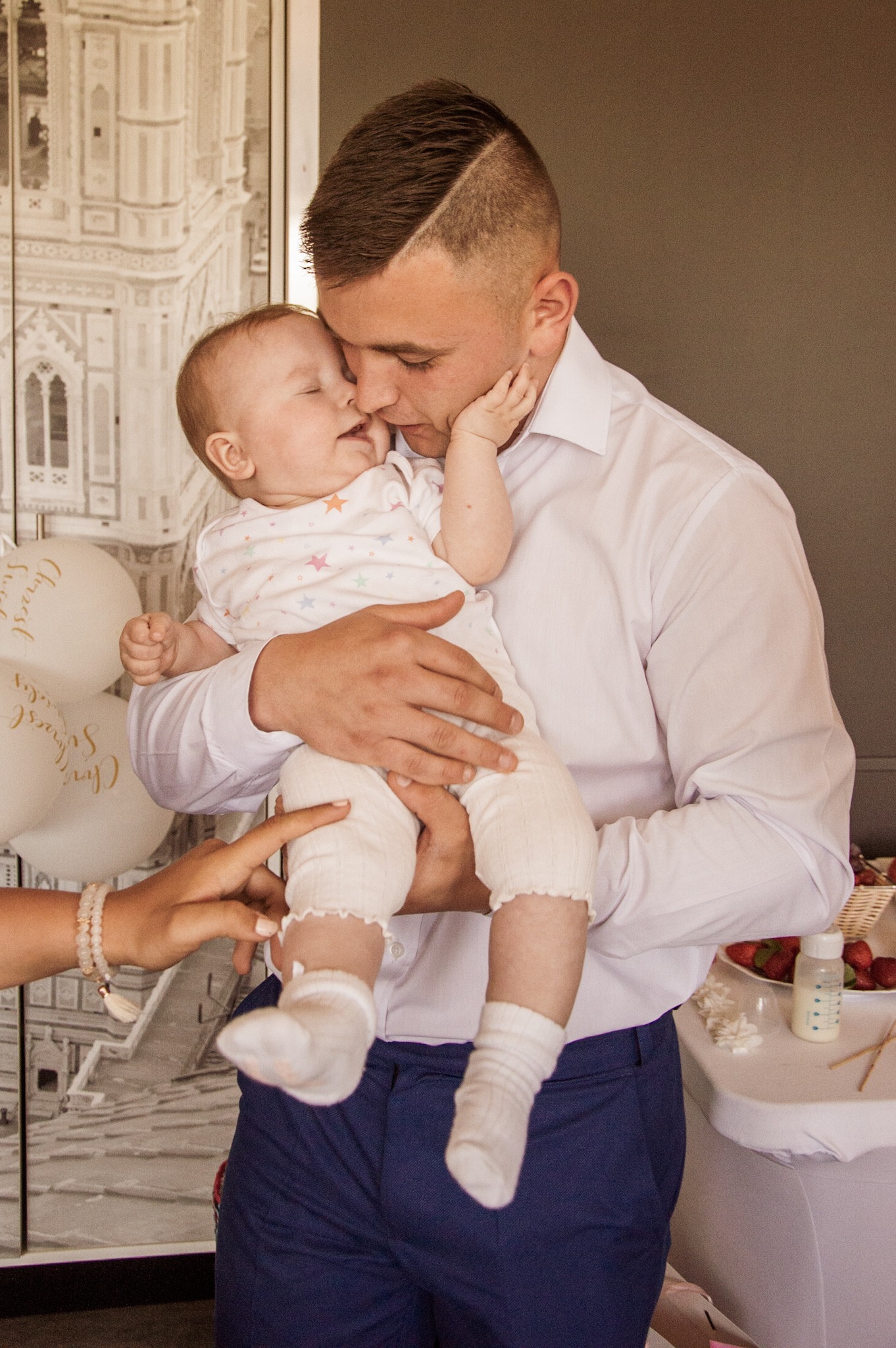 Fotograf eventowy uchwycił moment czułości między ojcem a dzieckiem podczas uroczystości; ojciec w białej koszuli i granatowych spodniach trzyma niemowlę w białym ubranku w gwiazdki na tle...