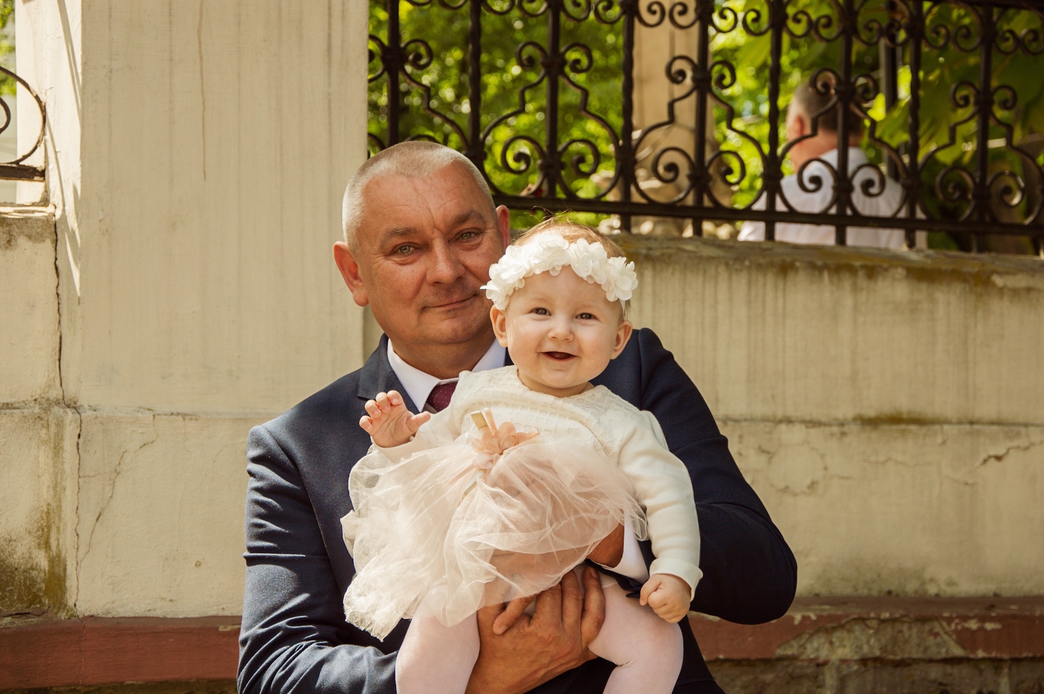 Uśmiechnięty mężczyzna w garniturze trzyma na rękach małą dziewczynkę w białej sukience i wianku na głowie, w tle ozdobna metalowa barierka i zieleń.
