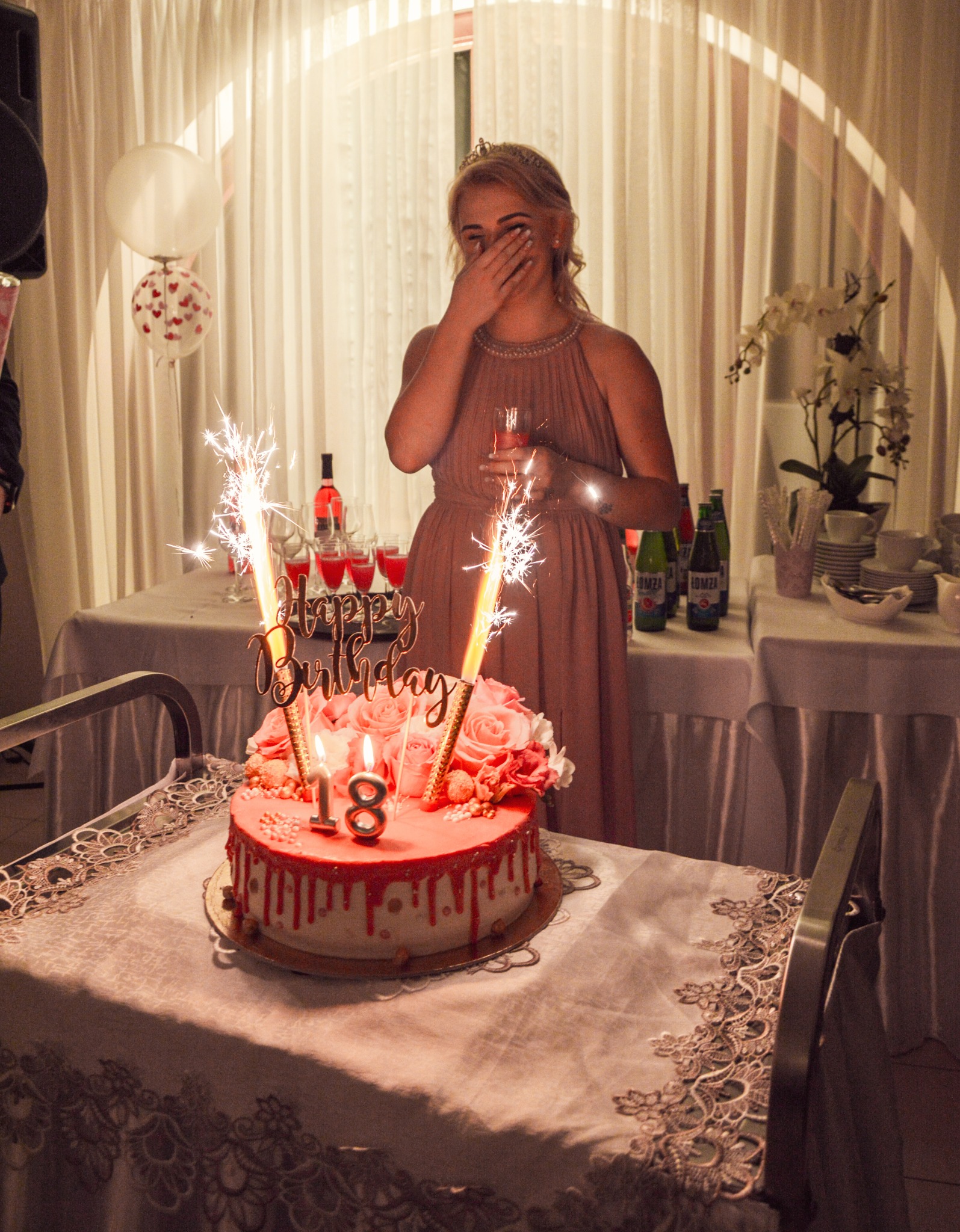 Uśmiechnięta blondynka z koroną zasłania twarz, trzymając kieliszek w dłoni, w tle tort urodzinowy z fontannami iskier na stole przykrytym koronkowym obrusem, balony i napoje