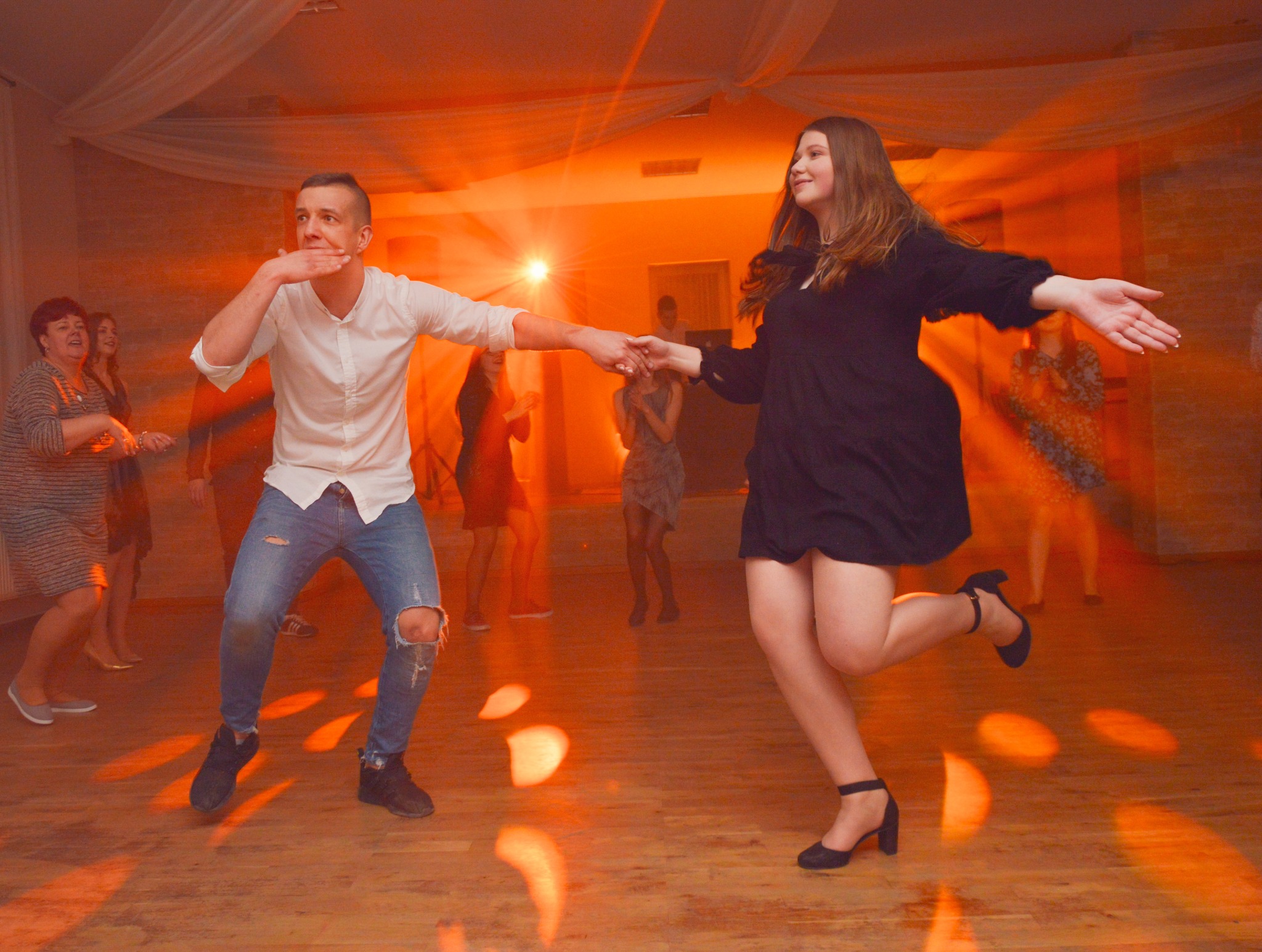 Para trzymająca się za ręce tańczy na parkiecie podczas imprezy, oświetlona pomarańczowym światłem, z innymi gośćmi w tle.