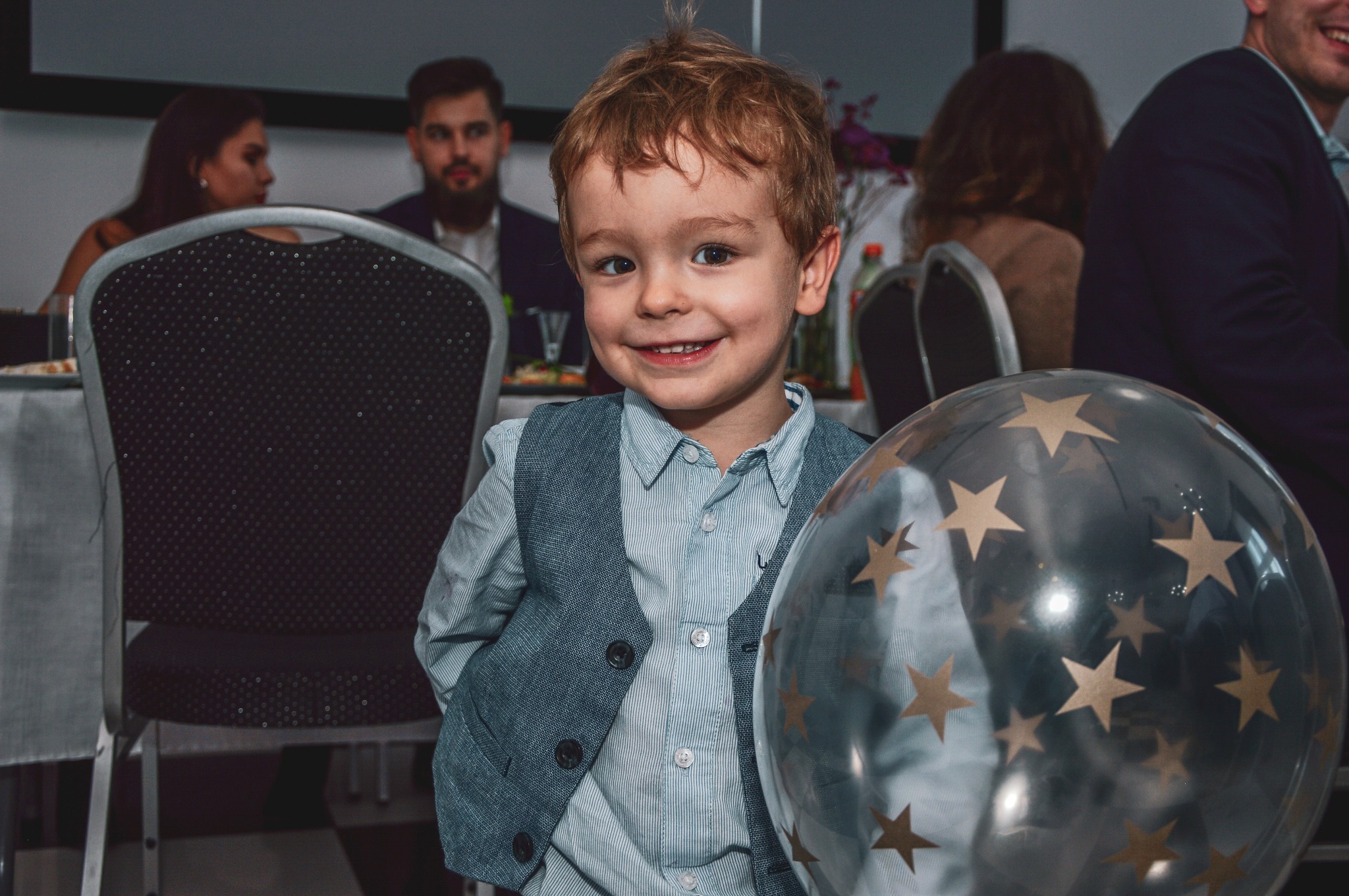 Uśmiechnięty chłopiec w eleganckiej kamizelce i koszuli, trzymający balon z gwiazdami na tle sali bankietowej z gośćmi.