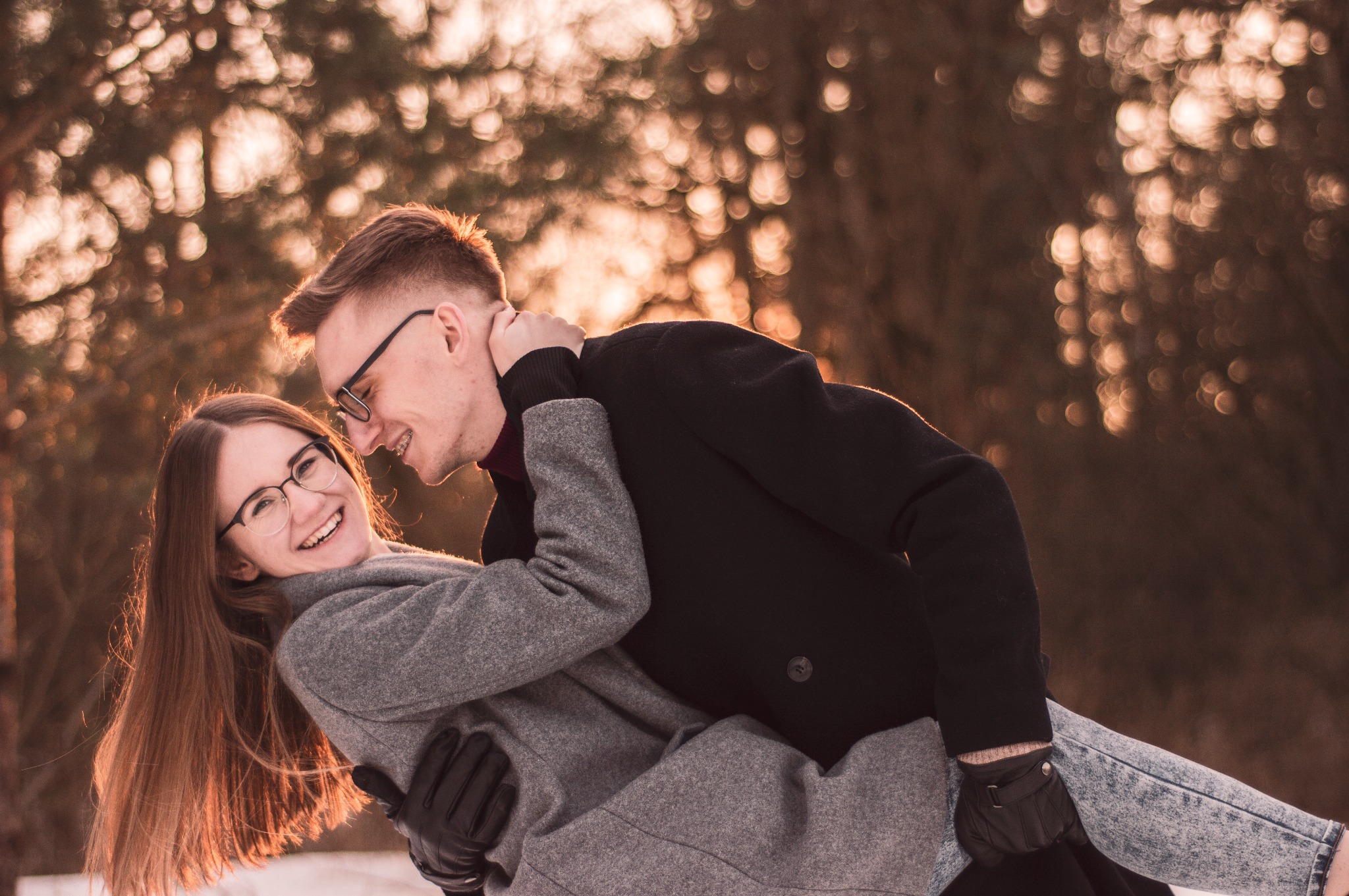 Uśmiechnięta para w okularach, on w czarnym płaszczu, ona w szarym, w objęciach na tle rozmytego zimowego krajobrazu o zachodzie słońca.