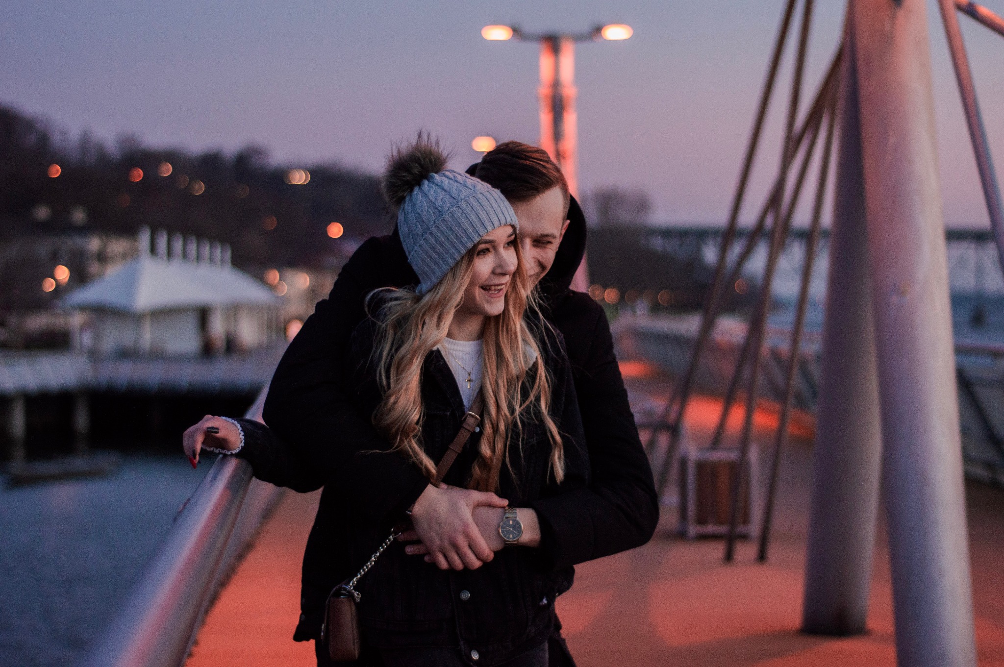 Para obejmująca się na moście o zmierzchu, kobieta w szarej czapce z pomponem i złoty naszyjnik krzyżyk, mężczyzna w czarnej kurtce z zegarkiem na nadgarstku.