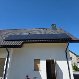 Eldan - Cztery czarne panele fotowoltaiczne zamontowane na ciemnoszarym dachu z dachówki, na tle błękitnego nieba. Obok biała elewacja domu z widoczną drabiną.