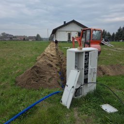 Eldan - Otwarta skrzynka przyłączeniowa z widocznymi zabezpieczeniami, niebieska rura osłonowa i świeżo wykopany rów prowadzący do budowanego domu, w tle mini-koparka.