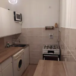 Mała kuchnia z białymi szafkami, drewnianym blatem, pralką pod blatem, kuchenką gazową, drewnianym stołem i płytkami w beżowym odcieniu.