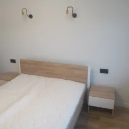 Nowoczesna sypialnia z łóżkiem z drewnianym zagłówkiem i białymi szafkami nocnymi, oświetlona dwoma czarnymi lampami ściennymi z żarówkami w stylu retro, na tle gładkiej, białej ściany.