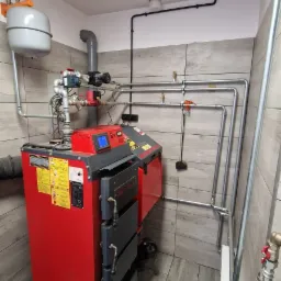 Czerwony piec centralnego ogrzewania z szarymi elementami, zamontowany w pomieszczeniu wyłożonym płytkami imitującymi drewno, widoczne rury instalacji, zbiornik wyrównawczy i czarne przewody...