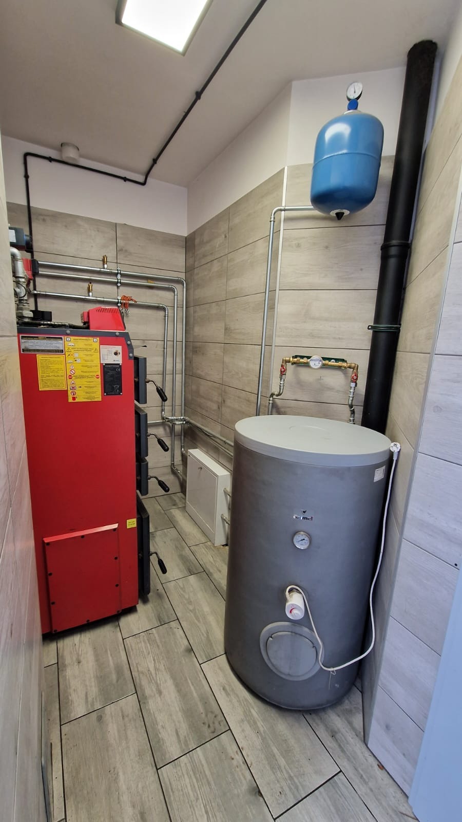 Czerwony piec na paliwo stałe, szary zasobnik ciepłej wody i niebieskie naczynie przeponowe w kotłowni wyłożonej płytkami imitującymi drewno.