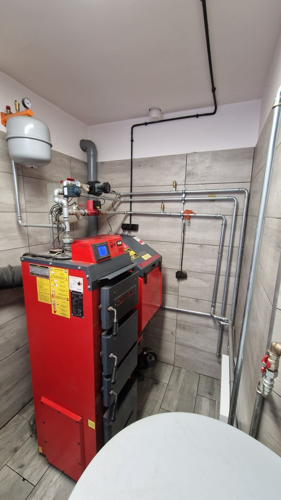Czerwony piec centralnego ogrzewania z szarymi elementami, zamontowany w pomieszczeniu wyłożonym płytkami imitującymi drewno, widoczne rury instalacji, zbiornik wyrównawczy i czarne przewody...