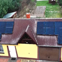 Instalacja paneli słonecznych na dachu domu jednorodzinnego z brązową blachodachówką, widok z góry na częściowo ośnieżony dach i otoczenie.