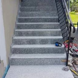 Zewnętrzne schody wejściowe wyłożone szarą, nakrapianą płytką, z czarną metalową balustradą po prawej stronie, widoczne narzędzia i akcesoria budowlane sugerują trwające prace.