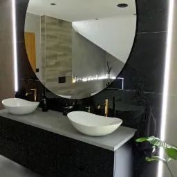 Nowoczesna łazienka z dwoma białymi umywalkami nablatowymi, złotymi bateriami, czarną szafką pod umywalką i okrągłym lustrem podświetlonym paskami LED po bokach, odbijającym fragment drugiej łazienki.
