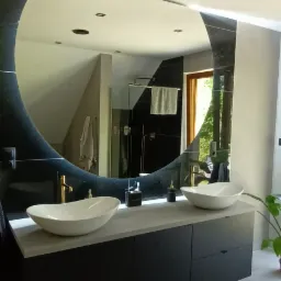 Nowoczesna łazienka z dwoma owalnymi umywalkami nablatowymi, czarną szafką pod umywalką, dużym okrągłym lustrem z podświetleniem LED i widokiem na kabinę prysznicową w tle.