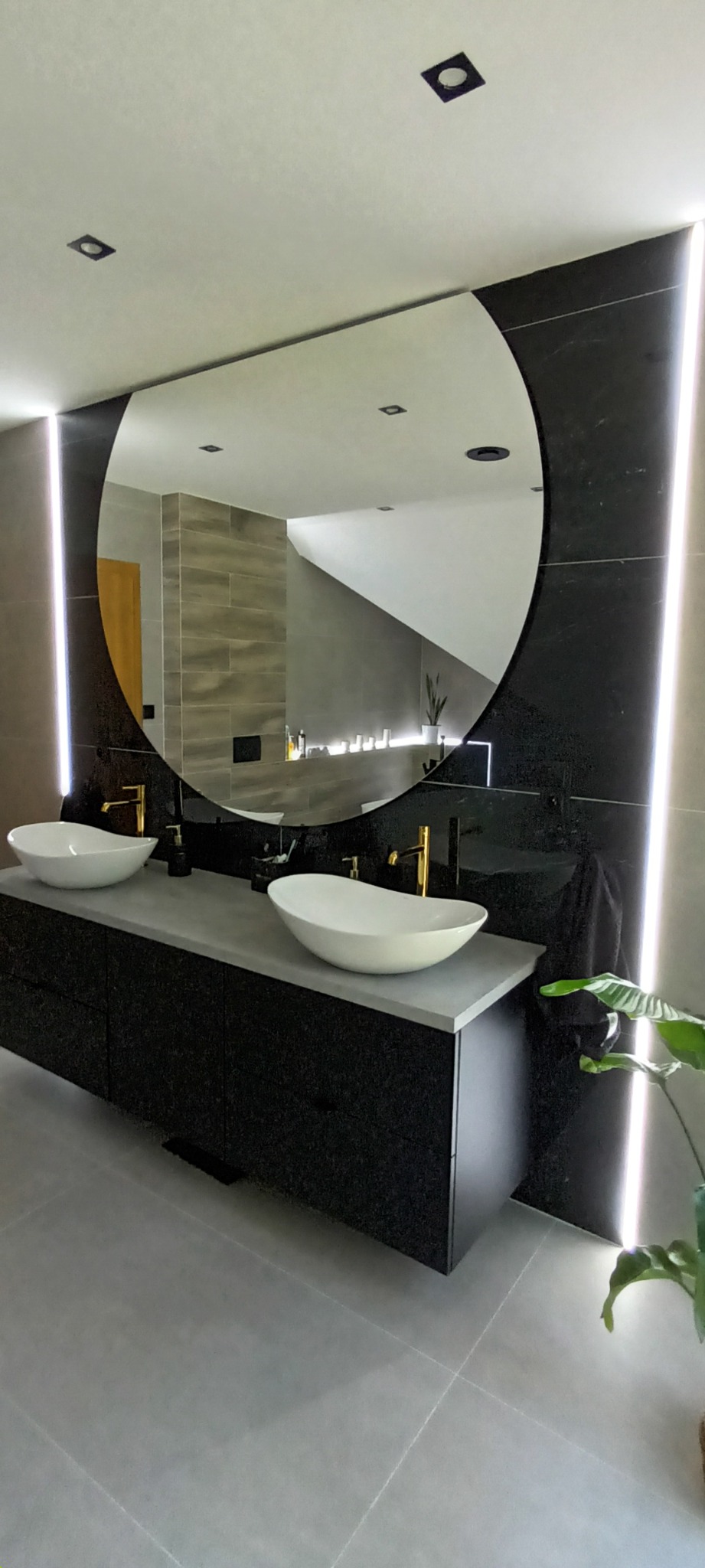 Nowoczesna łazienka z dwoma białymi umywalkami nablatowymi, złotymi bateriami, czarną szafką pod umywalką i okrągłym lustrem podświetlonym paskami LED po bokach, odbijającym fragment drugiej łazienki.