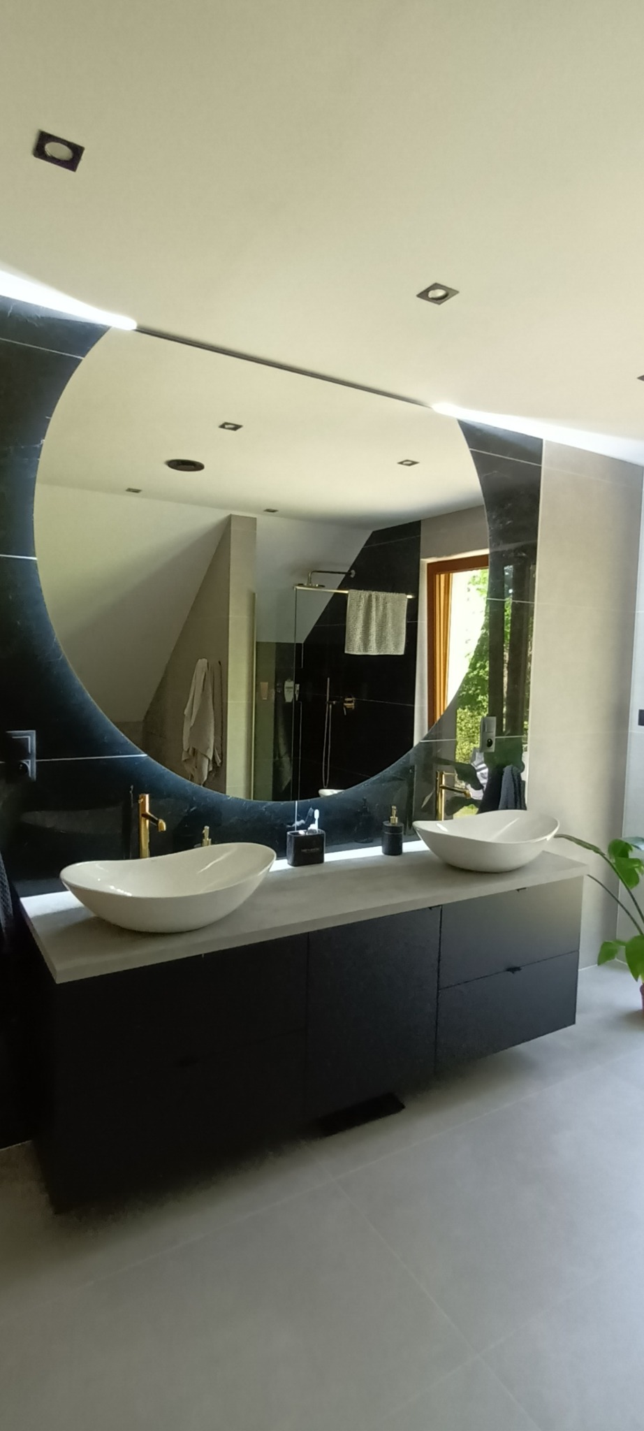 Nowoczesna łazienka z dwoma owalnymi umywalkami nablatowymi, czarną szafką pod umywalką, dużym okrągłym lustrem z podświetleniem LED i widokiem na kabinę prysznicową w tle.