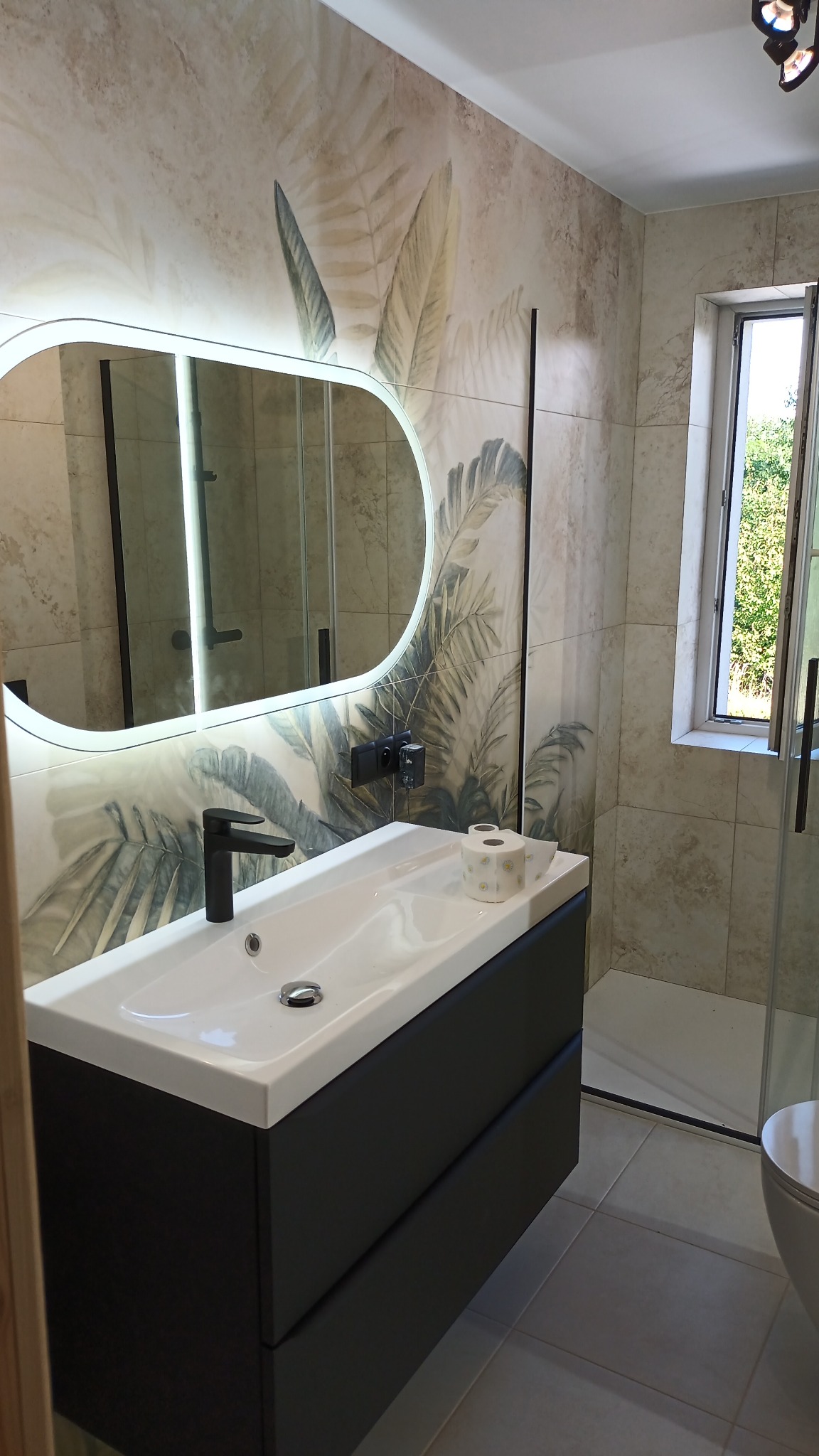 Nowoczesna łazienka z umywalką nablatową, czarną szafką i podświetlanym lustrem owalnym. Na ścianie płytki z motywem liści. Widoczny fragment kabiny prysznicowej.