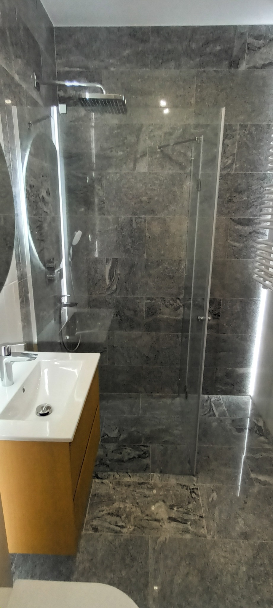 Nowoczesna łazienka z szarymi płytkami imitującymi kamień, kabiną prysznicową typu walk-in i umywalką z szafką w kolorze drewna. Widoczne oświetlenie LED.