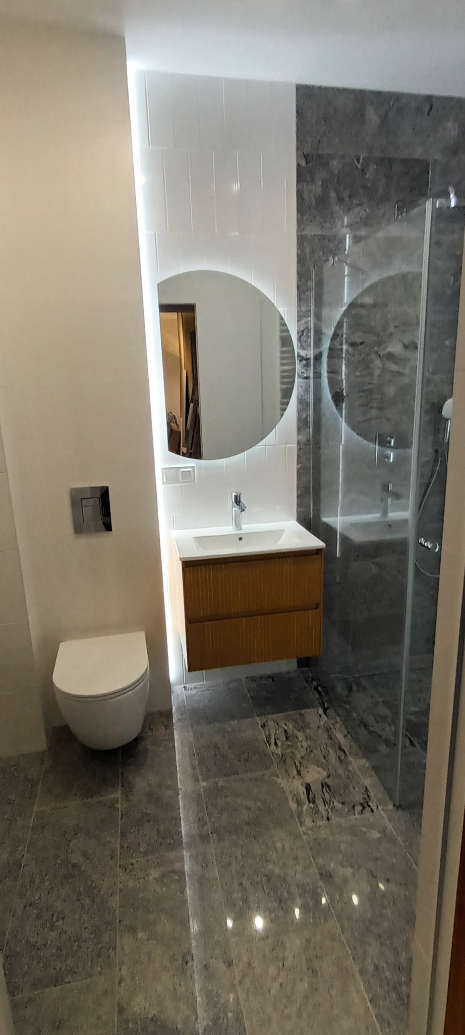 Nowoczesna łazienka z owalnym lustrem podświetlanym LED, drewnianą szafką pod umywalką i kabiną prysznicową z szarymi płytkami imitującymi marmur.