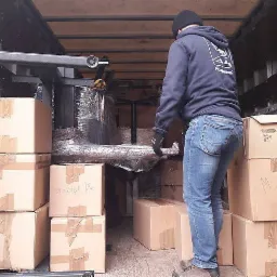 Pracownik w czapce i bluzie z kapturem, w jeansach i rękawicach, układa kartony wewnątrz furgonetki; widoczny fragment zapakowanego mebla zabezpieczonego folią stretch.