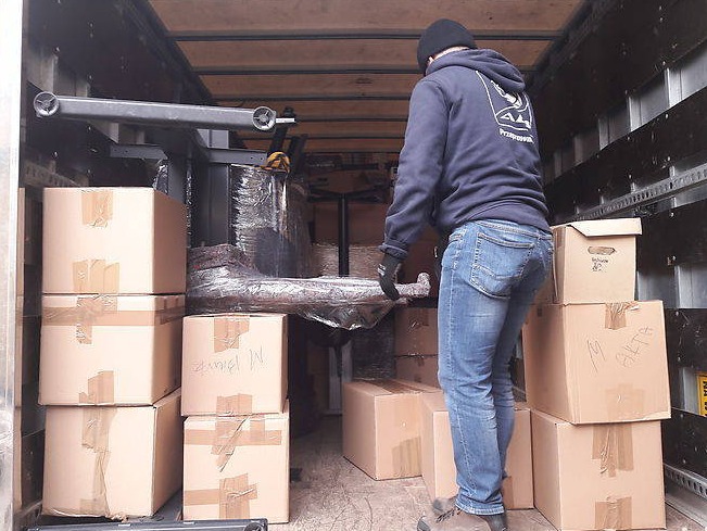 Pracownik w czapce i bluzie z kapturem, w jeansach i rękawicach, układa kartony wewnątrz furgonetki; widoczny fragment zapakowanego mebla zabezpieczonego folią stretch.