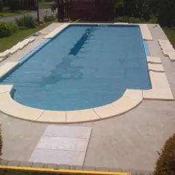 Basen z pokrywą, wykończony jasnymi płytkami, otoczony zielenią, widok z góry na ogród.