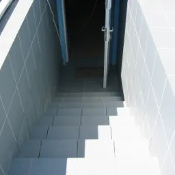 Kafelkowane schody w dół, prowadzące do otwartych drzwi z metalową kratką wentylacyjną na podłodze, wykończone jasnoszarymi płytkami z białymi fugami.