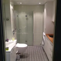 Wąska łazienka z kabiną prysznicową, toaletą, umywalką na szafce, pralką i szafkami z drewnianym blatem, wykończona jasnymi płytkami i szarą podłogą w kratkę, widok zza futryny.