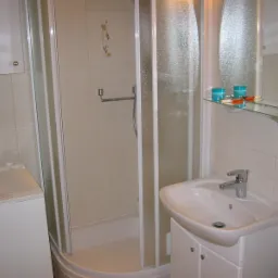 Jasna łazienka z kabiną prysznicową, umywalką z szafką, pralką i szklaną półką z kosmetykami, wykończona płytkami w odcieniach beżu.