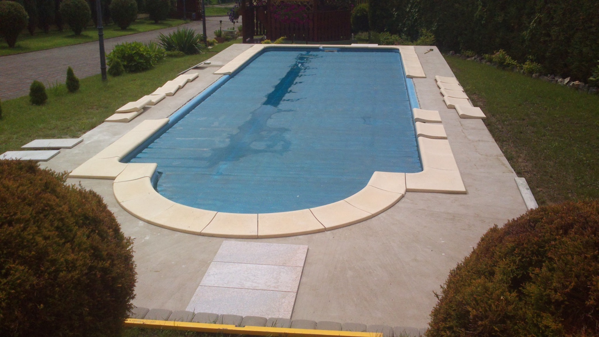 Basen z pokrywą, wykończony jasnymi płytkami, otoczony zielenią, widok z góry na ogród.