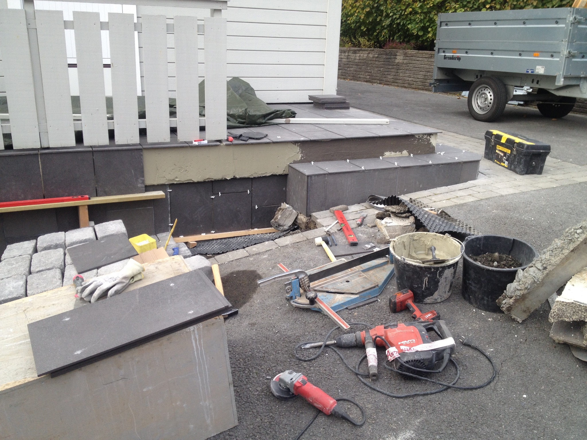 Montaż ciemnoszarych płytek na schodach zewnętrznych; widoczne narzędzia glazurnicze, kostka brukowa i przyczepa.