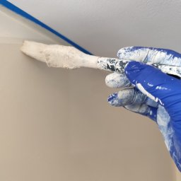 POMIMO Marcin Krzycki - Malowanie ściany pędzlem przy suficie, zabezpieczonym niebieską taśmą. Ręka w niebieskiej rękawicy ochronnej. Widoczne ślady farby na pędzlu i rękawicy.