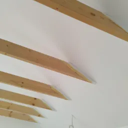 Biały sufit z drewnianymi belkami zabezpieczonymi taśmą malarską przed malowaniem, widoczny wystający przewód elektryczny.