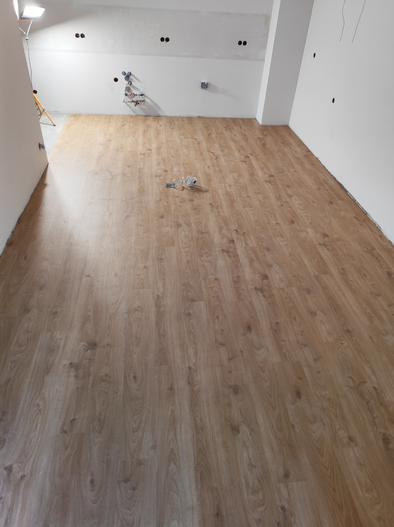 Jasnobrązowa podłoga laminowana w pustym pokoju z białymi ścianami i widocznymi instalacjami elektrycznymi/hydraulicznymi. W rogu widoczna drabina i lampa budowlana.