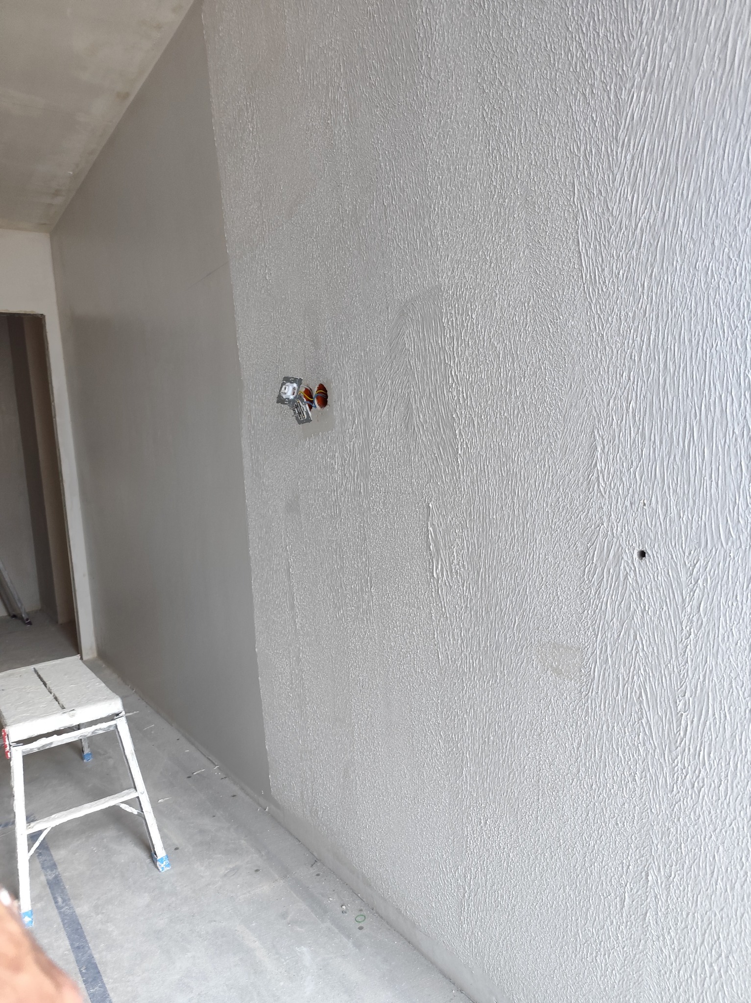Szara ściana z fakturą po nałożeniu gładzi, widoczne puszki elektryczne bez osprzętu i drabina malarska. Teksturowana powierzchnia ściany.