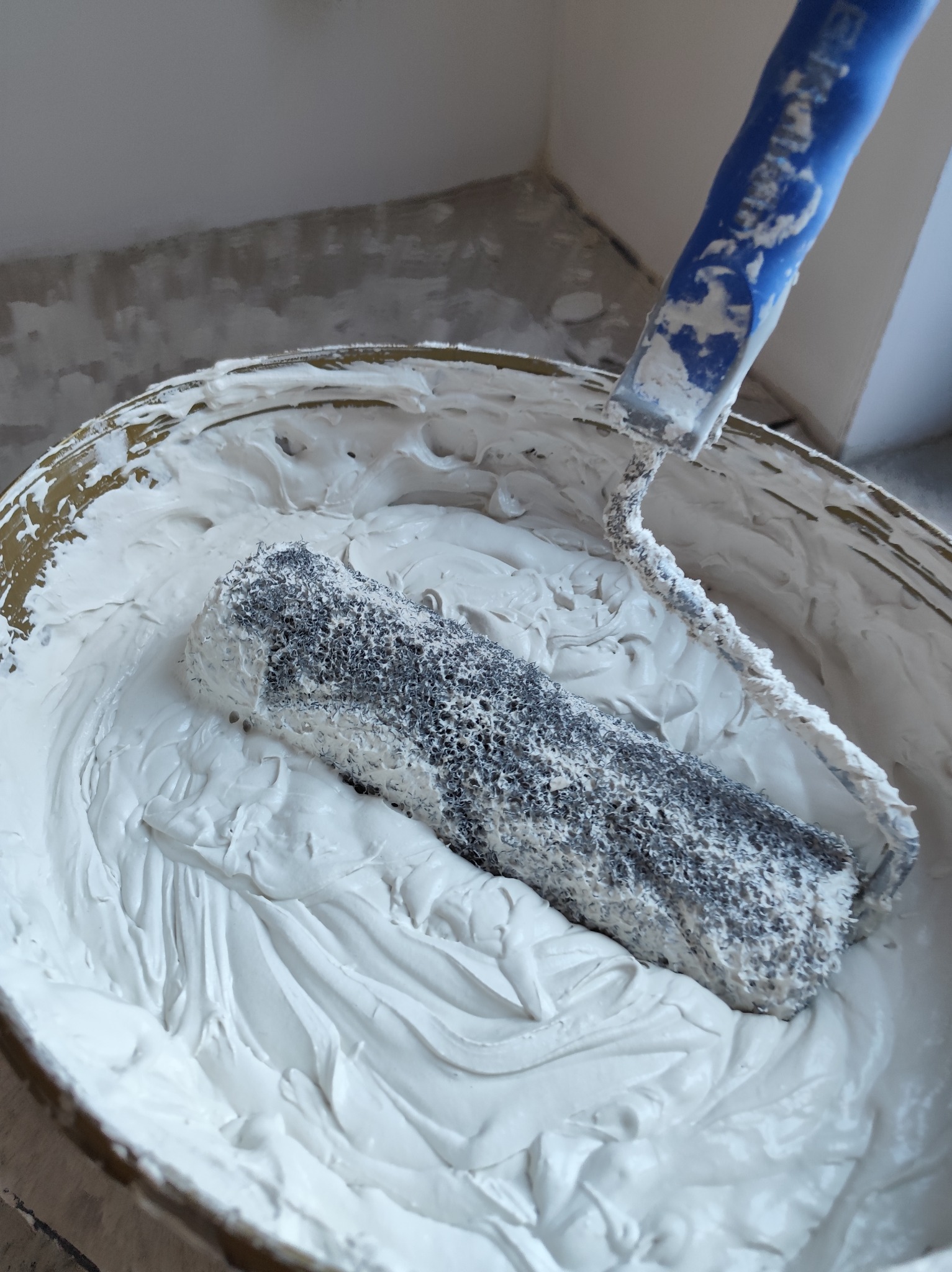 Wałek do malowania zanurzony w wiadrze z gładzią, widoczne ślady użycia, tło stanowią surowe ściany. Przygotowanie do prac wykończeniowych.