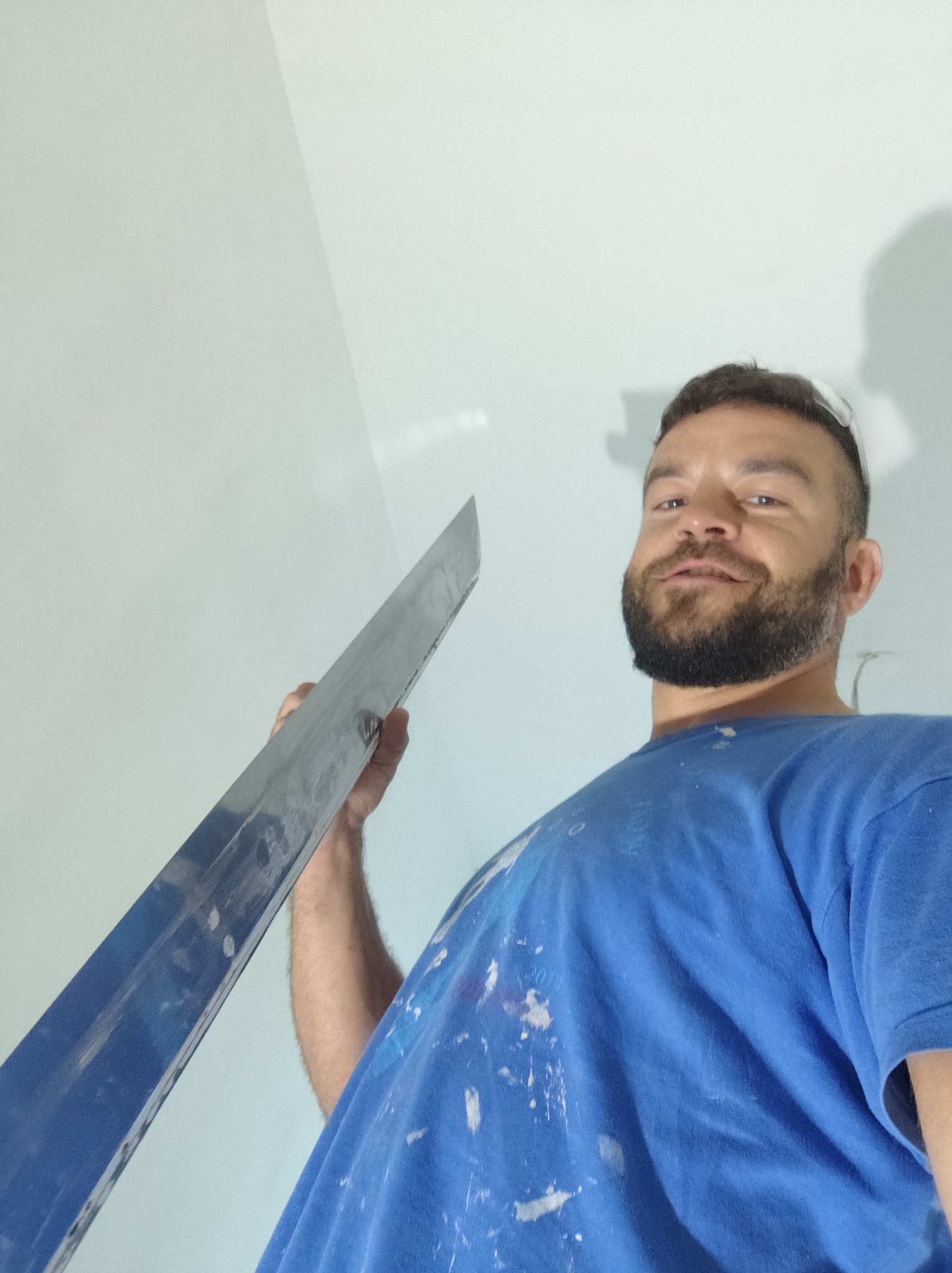 Mężczyzna z brodą, ubrany w niebieską koszulkę z plamami, trzyma w ręku długą szpachelkę na tle białej ściany. Ujęcie z dołu.