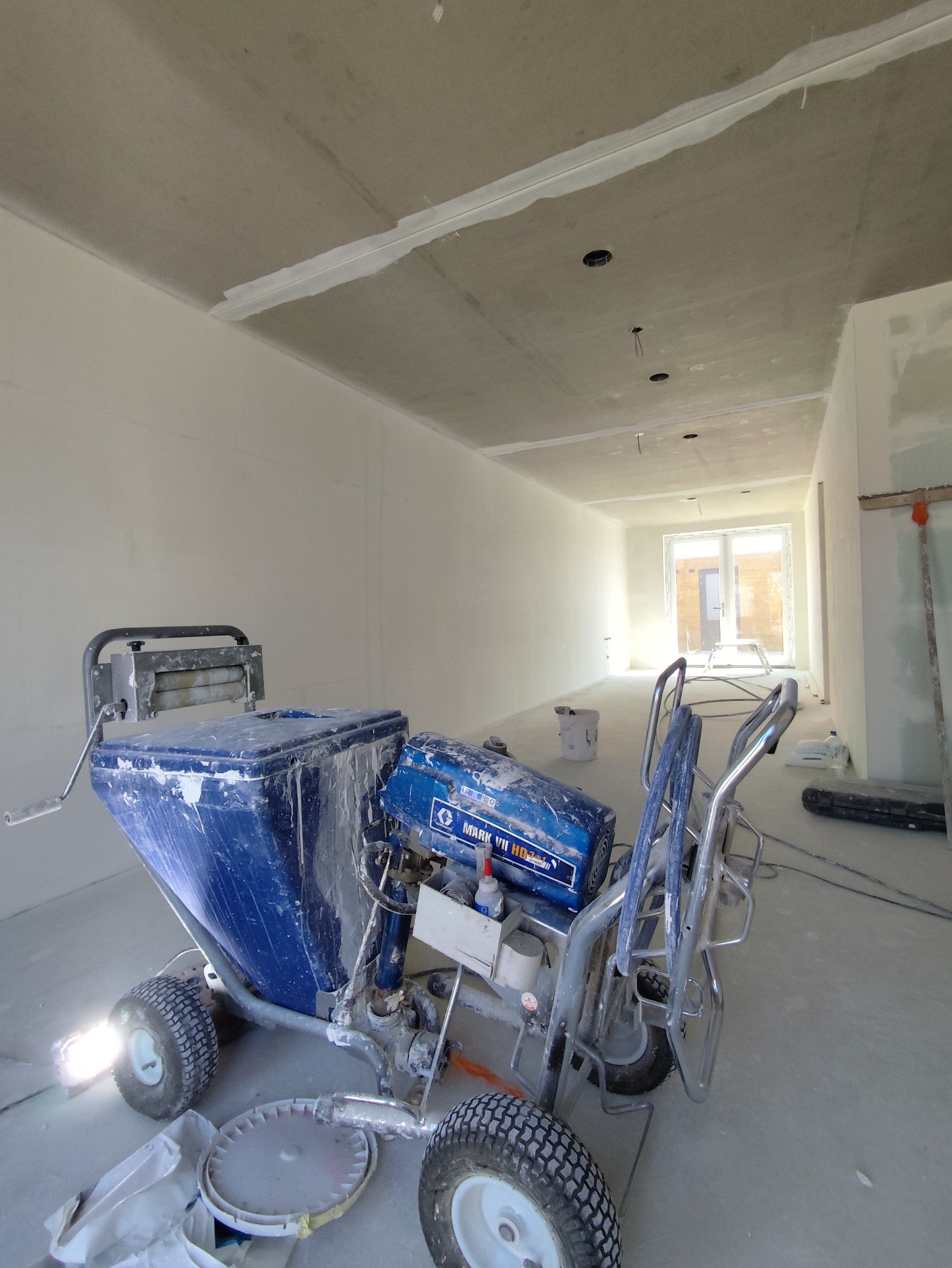 Wnętrze w trakcie prac wykończeniowych. Agregat malarski Graco na tle surowych ścian i sufitu z widocznymi łączeniami płyt. Otwory pod oświetlenie.