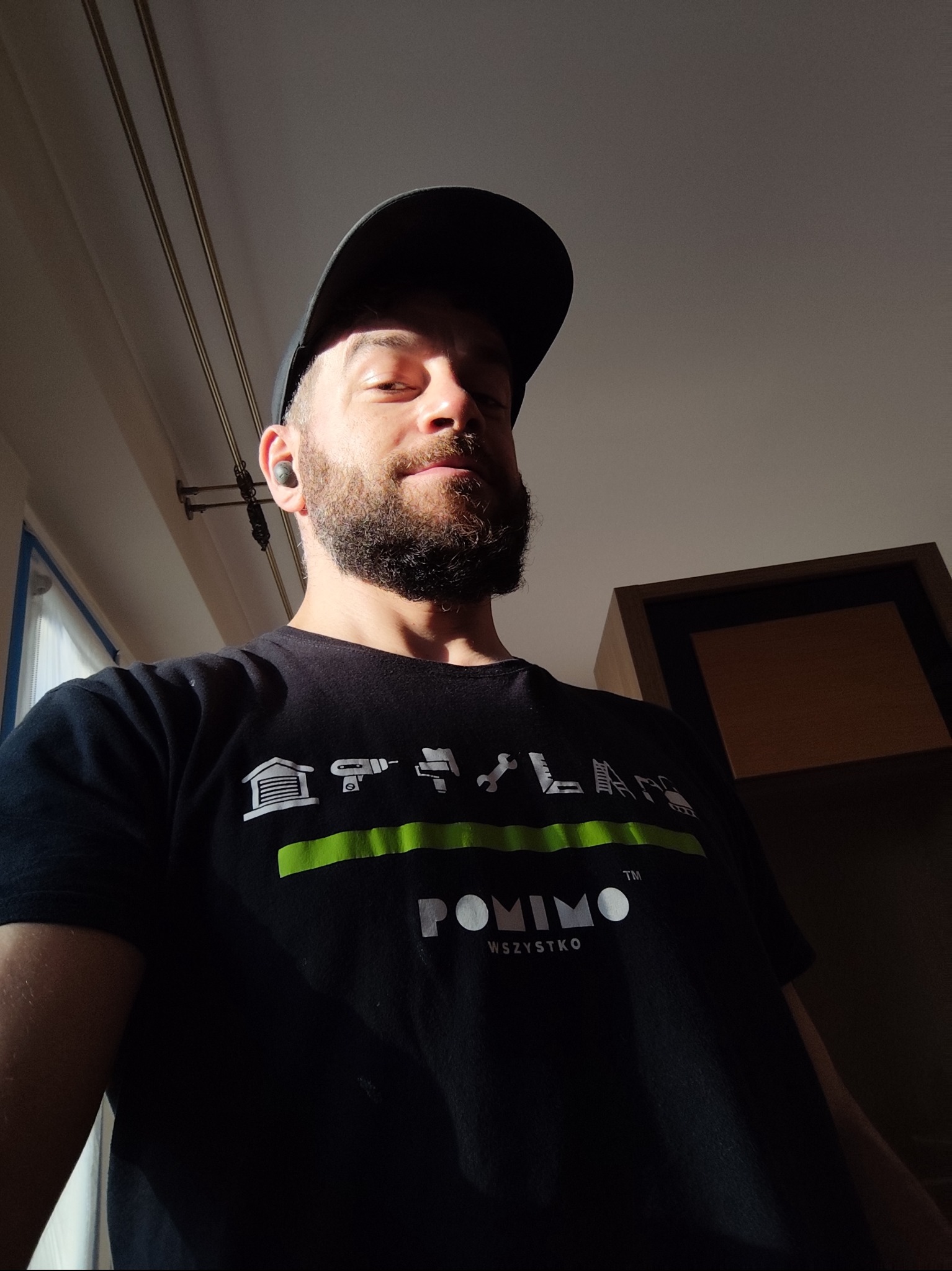 Mężczyzna z brodą w czarnej koszulce z grafiką narzędzi remontowych, w czapce z daszkiem, patrzy w dół, oświetlony promieniami słońca w pomieszczeniu.