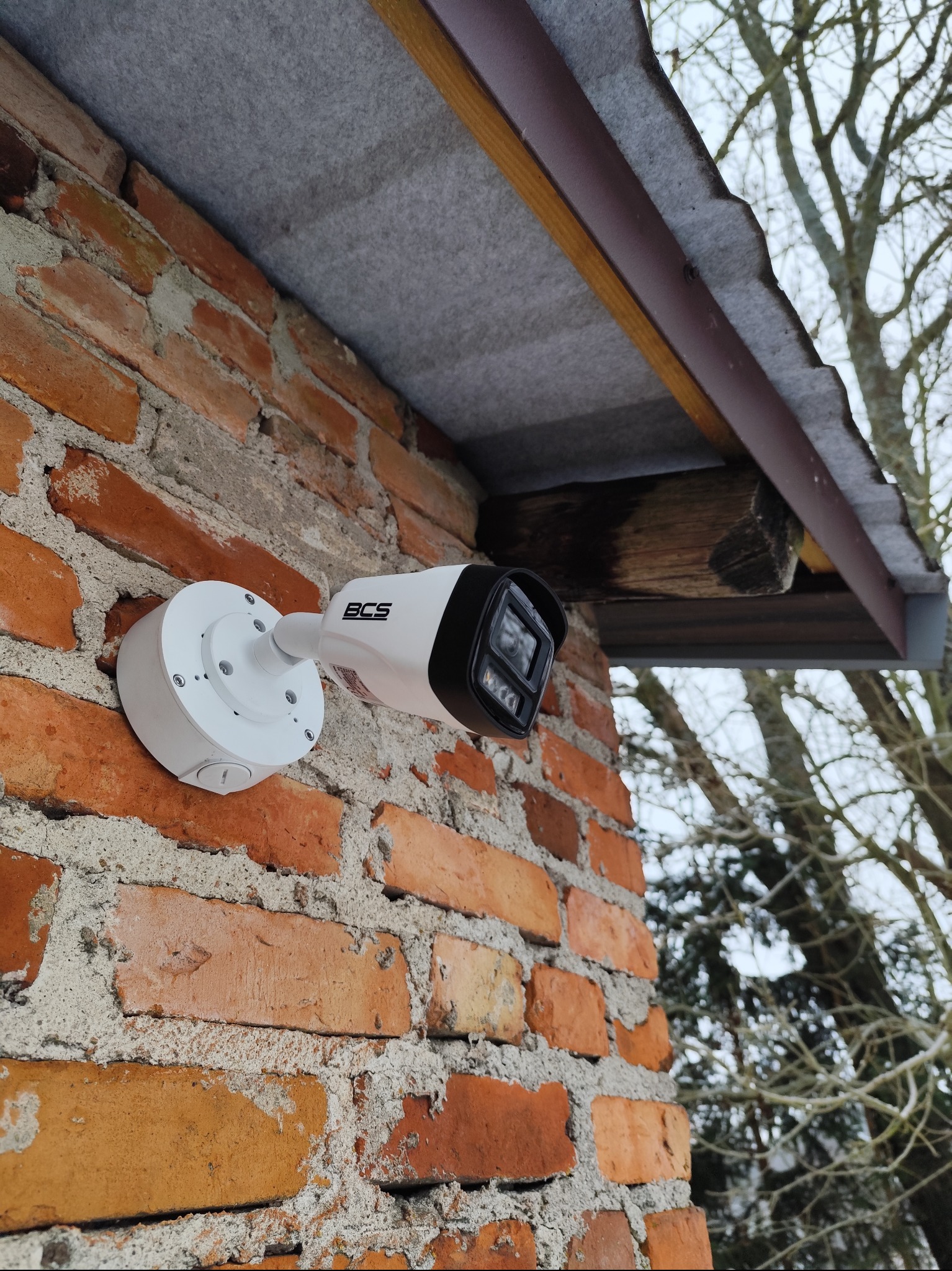 Kamera monitoringu BCS zamontowana na ceglanej ścianie budynku, widok z dołu, z fragmentem dachu i drzewem w tle. Widoczne śruby montażowe i logo producenta.