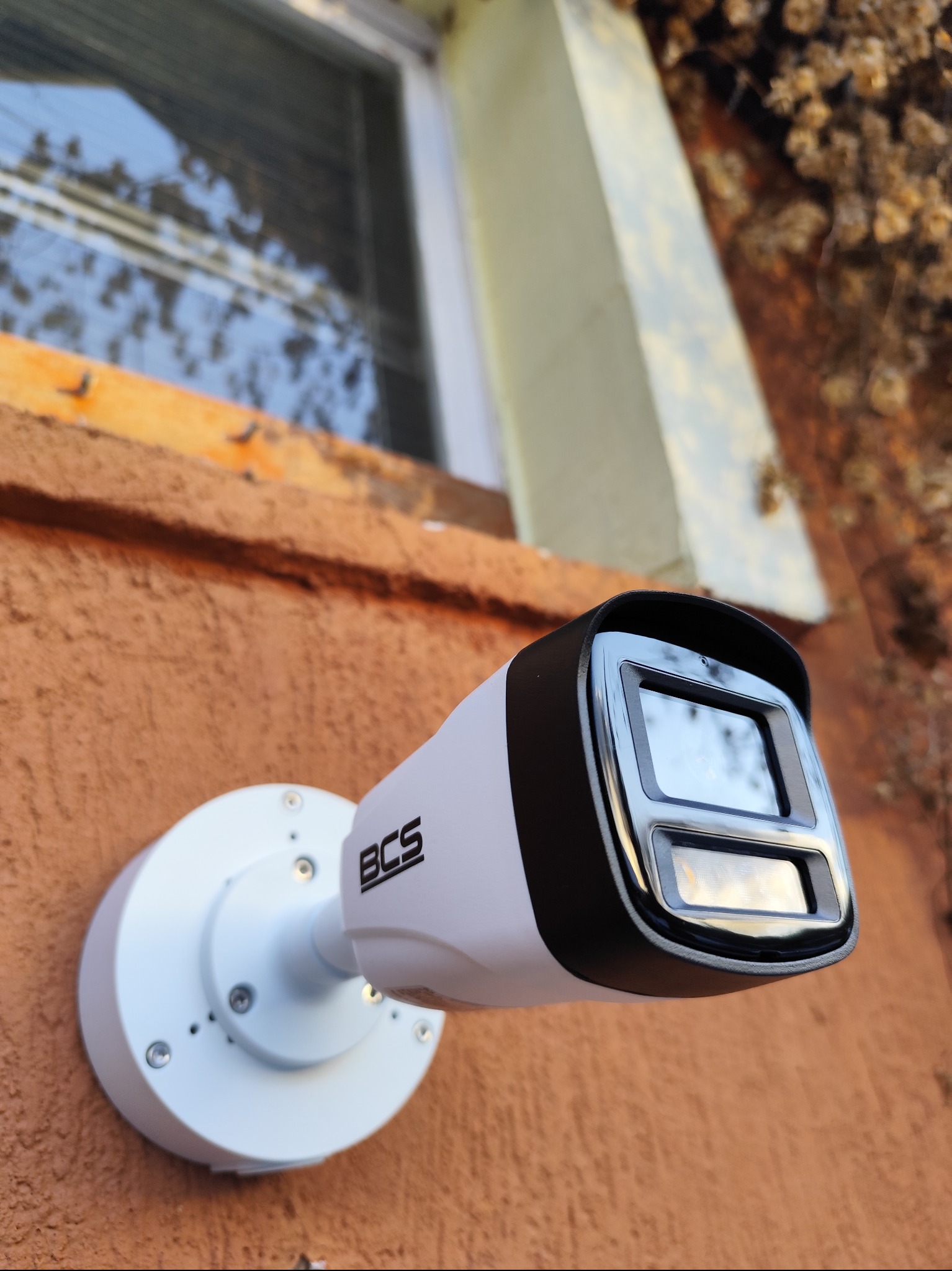 Zewnętrzna kamera monitoringu BCS, zamontowana na pomarańczowej ścianie budynku, widoczna z dołu pod kątem. W tle okno i fragment roślinności.