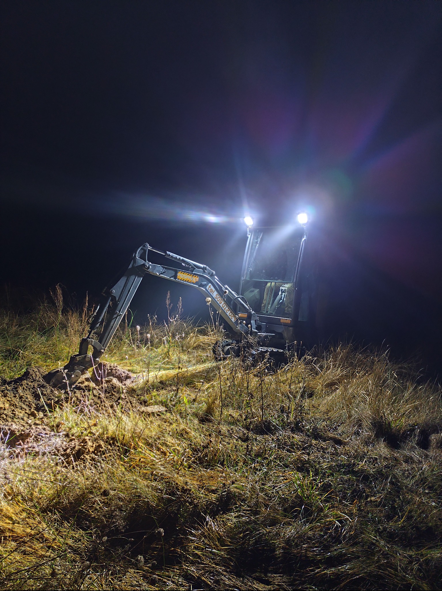Mini koparka w pracy nocą w Bydgoszczy. Oświetlona reflektorami, kopie ziemię na zarośniętym terenie. Widoczne trawy i grudki ziemi.