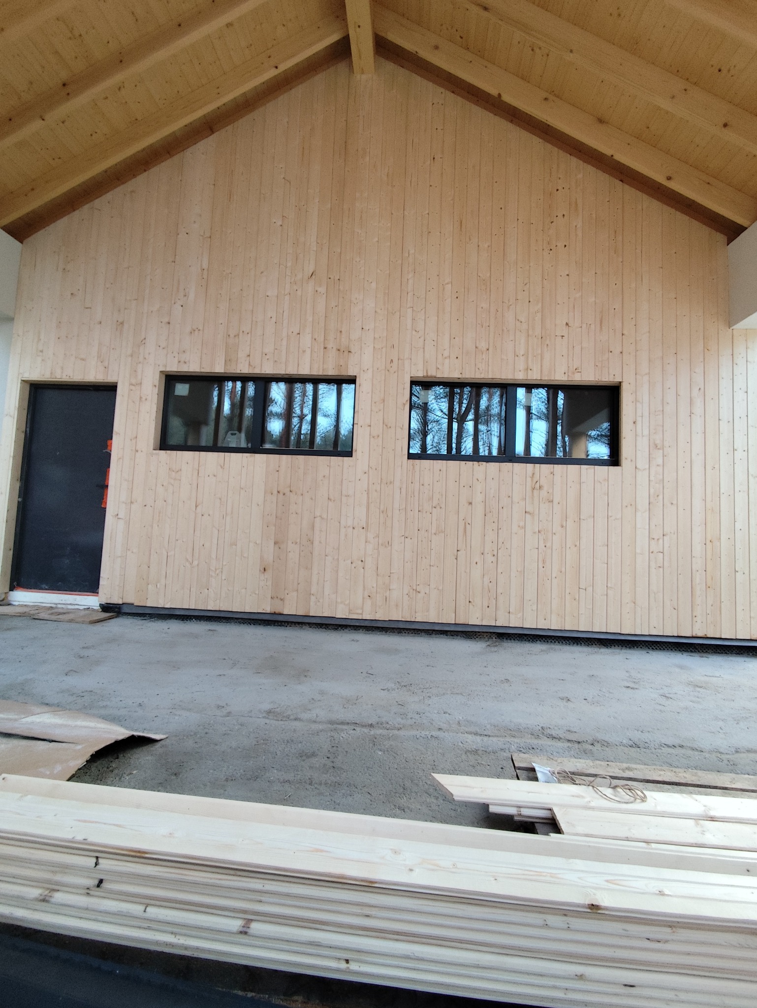 Wnętrze domu w trakcie wykańczania: drewniana boazeria na ścianach, dwa okna z czarnymi ramami odbijające drzewa, surowy beton na podłodze i stos desek.
