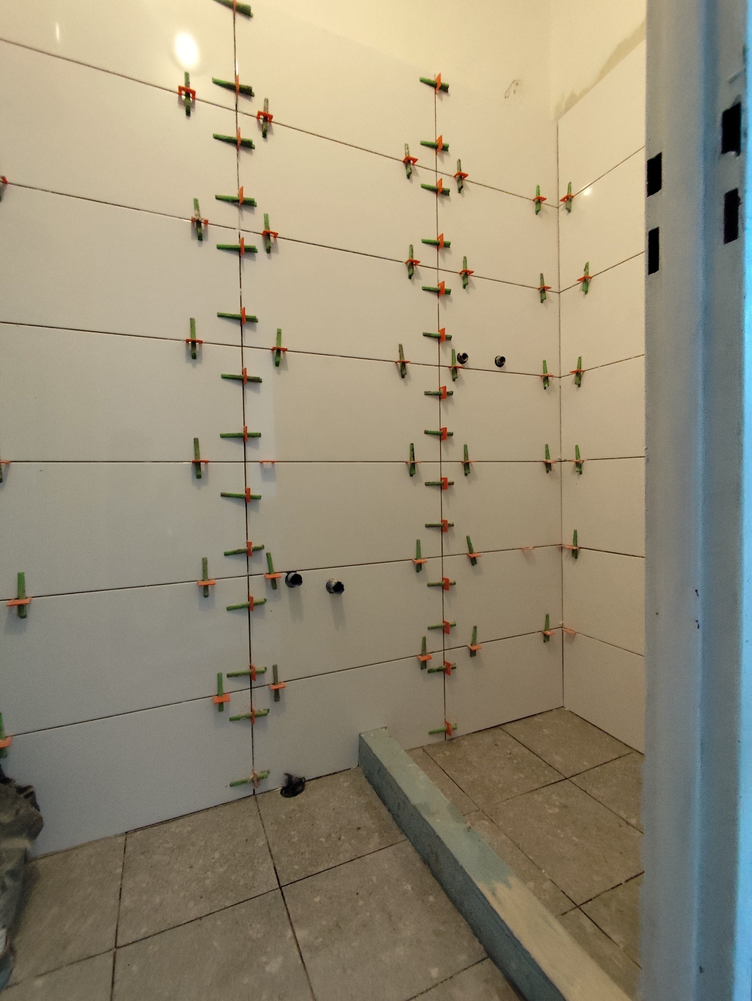 Świeżo ułożone, białe płytki w łazience, z systemem poziomowania płytek (klipsy i kliny) w kolorze zielono-pomarańczowym, widok na narożnik pomieszczenia.