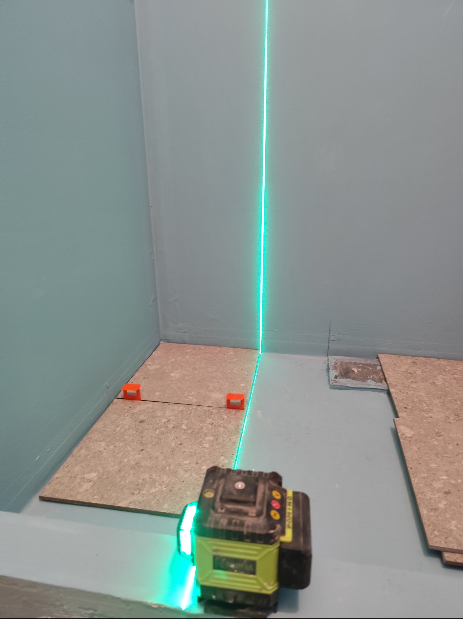 Układanie płytek podłogowych z użyciem poziomicy laserowej, widoczne płytki, krzyżyki dystansowe i zielona linia lasera na tle pomalowanej ściany.