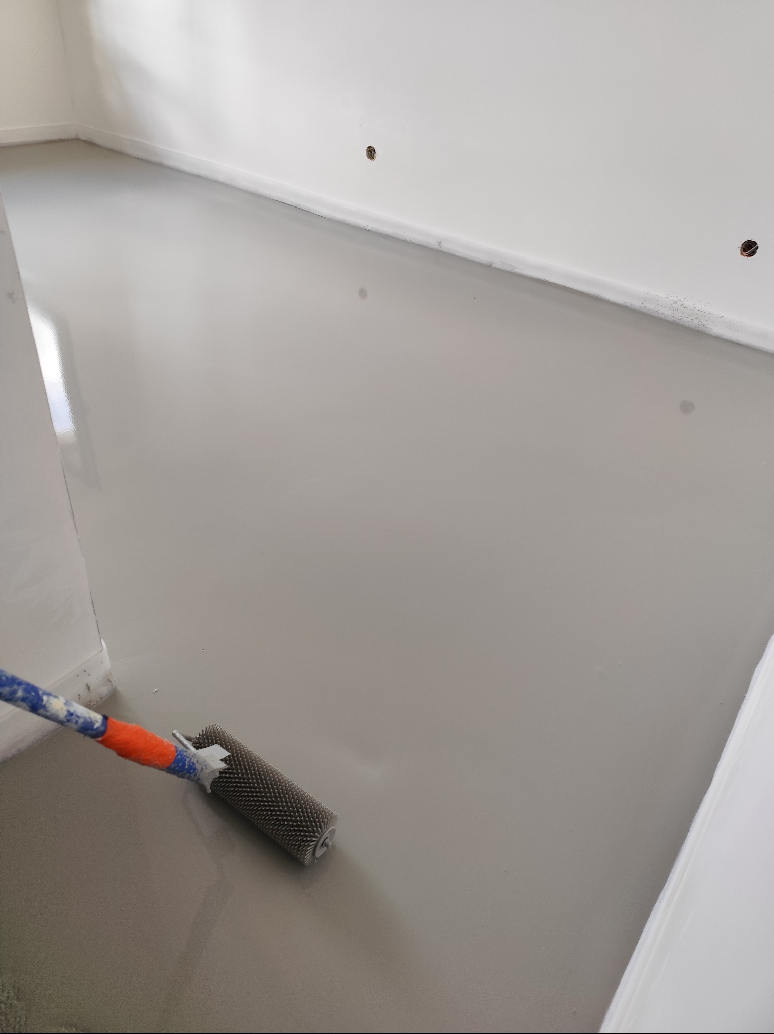 Szary, świeżo wylany samopoziomujący podkład podłogowy w jasnym pomieszczeniu. Widoczny wałek kolczasty do odpowietrzania wylewki.